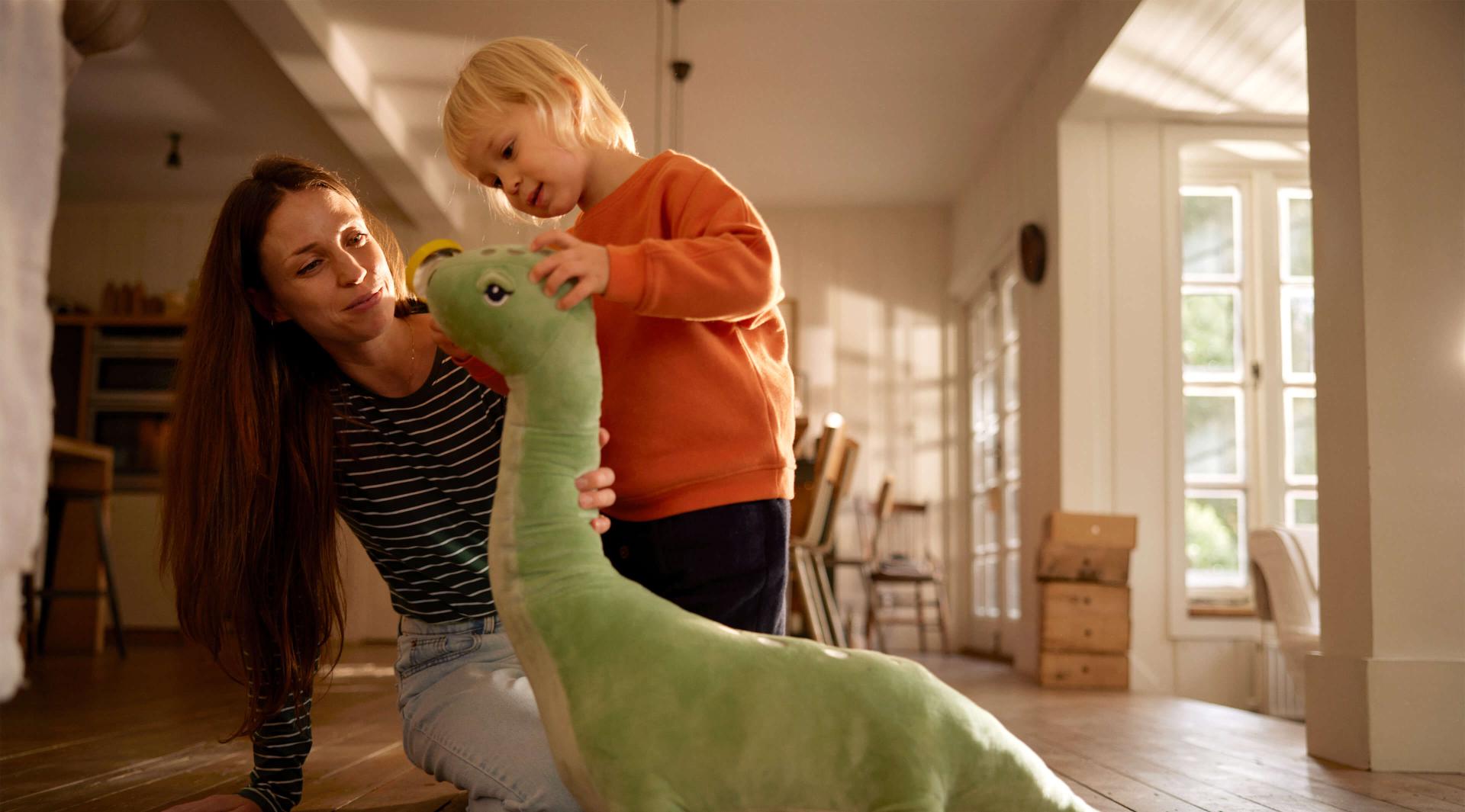 Frau und Kind spielen mit einem großen Stofftierdinosaurier.